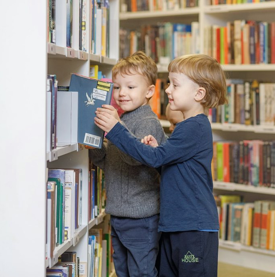 Du berniukai renkasi knygas bibliotekos lentynoje